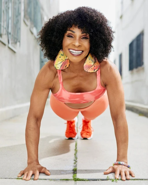An adult woman is doing a push-up in an outdoor setting with athletic wear and wearing decorative earrings with a smile on her face.