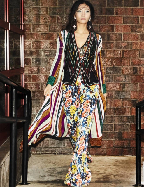 A long shot captures an Asian woman in a patterned floral dress and striped duster, standing in front of a brick wall near metal railings.
