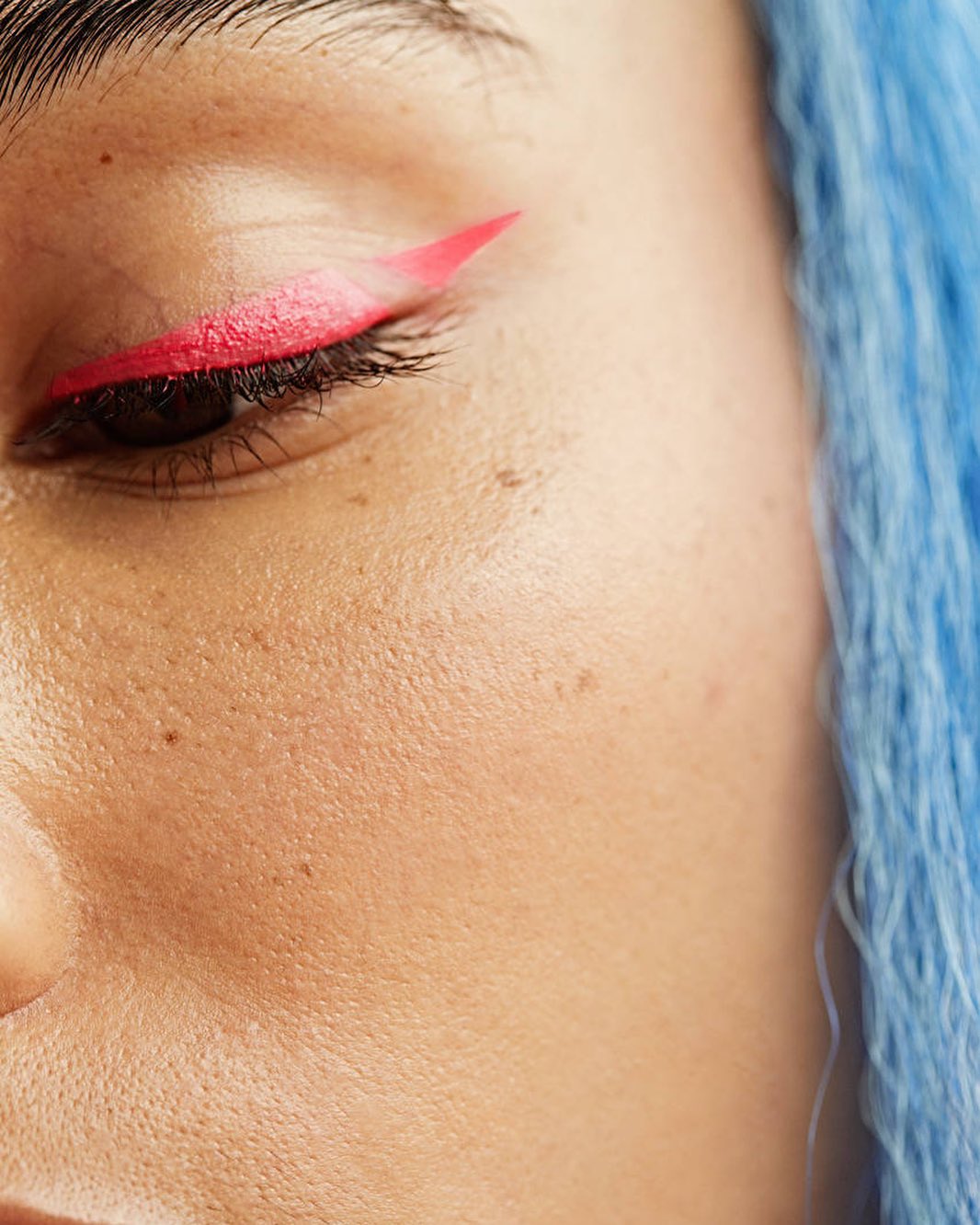 Close-up of a woman's face showcasing bold pink eyeliner in a winged design and blue hair, emphasizing the beauty and makeup elements.