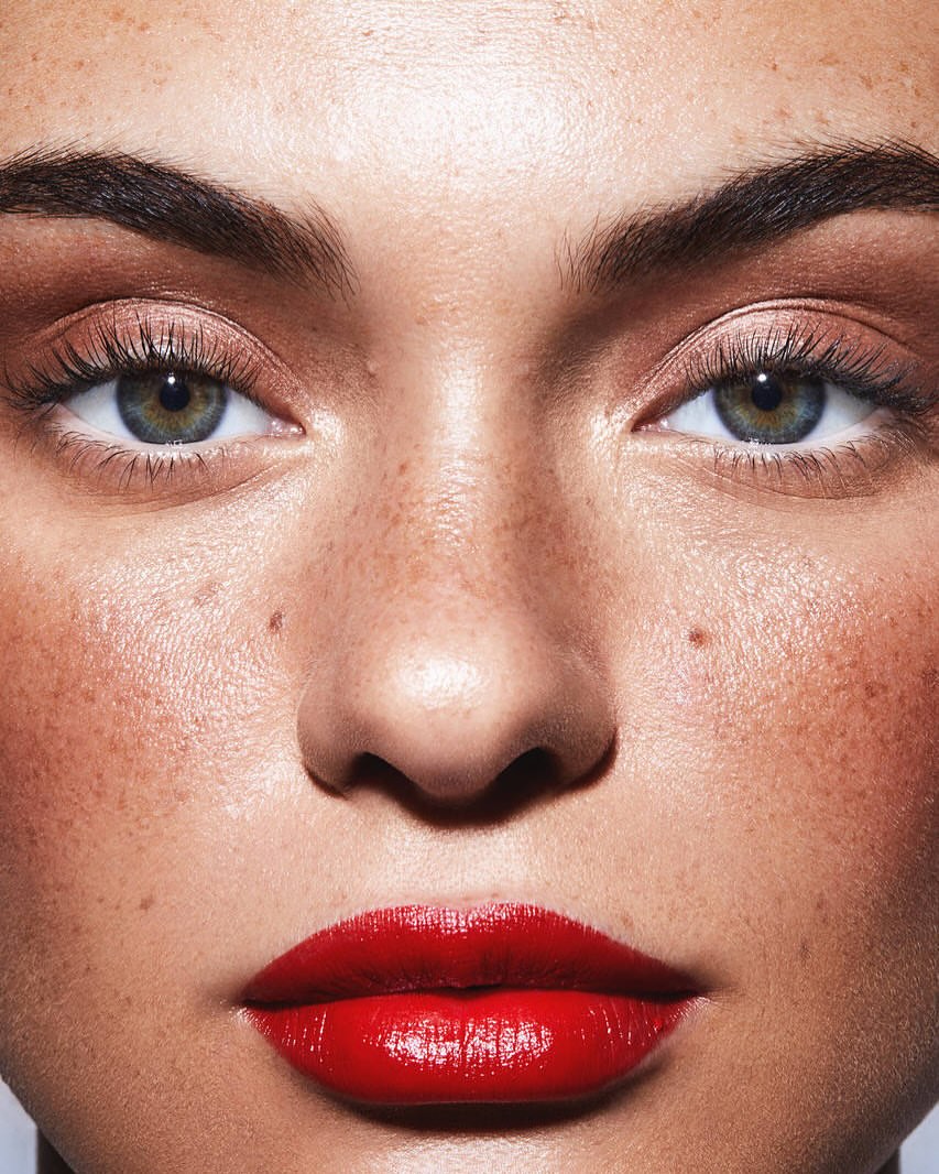 Close-up portrait of a fair-skinned woman with red lipstick, brown eye shadow, and freckles, focusing on her facial features and makeup.