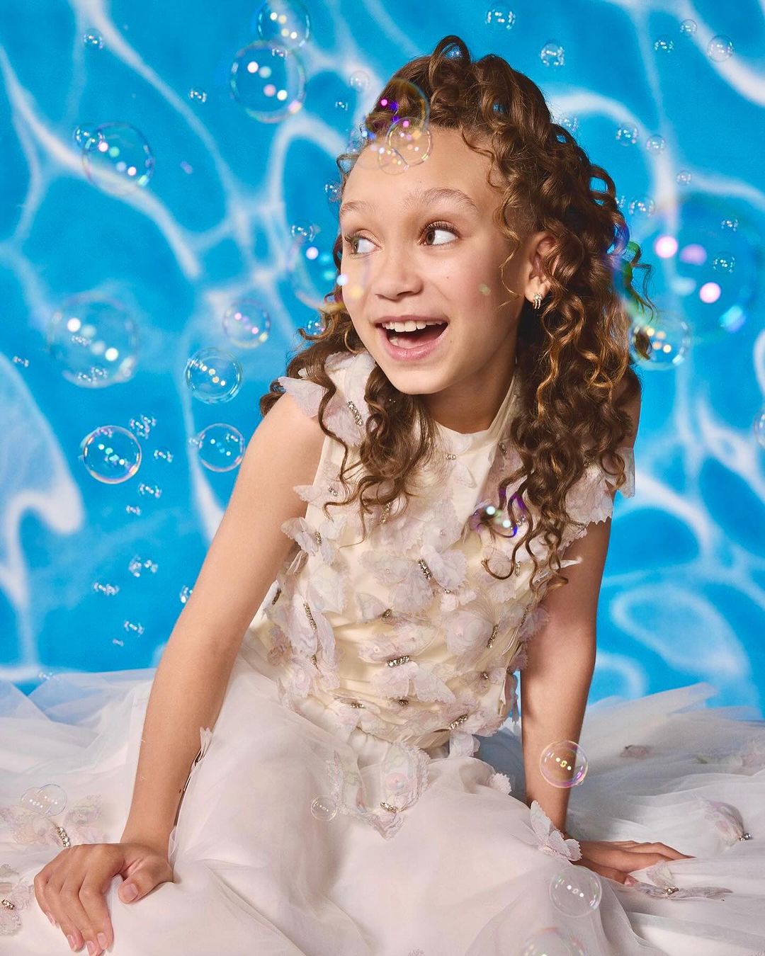 Smiling girl in a butterfly-embellished white dress poses in front of a water-patterned backdrop with floating soap bubbles around her.