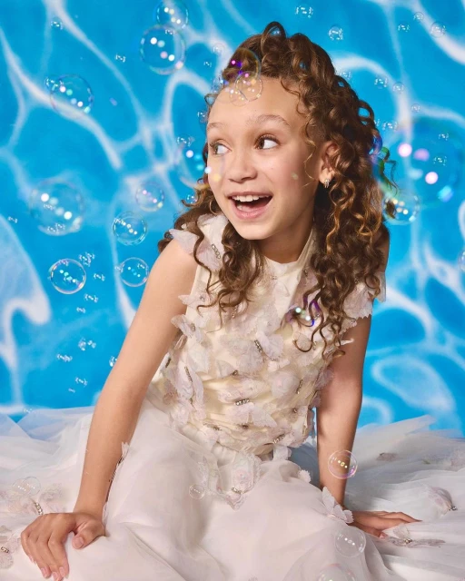 Smiling girl in a butterfly-embellished white dress poses in front of a water-patterned backdrop with floating soap bubbles around her.