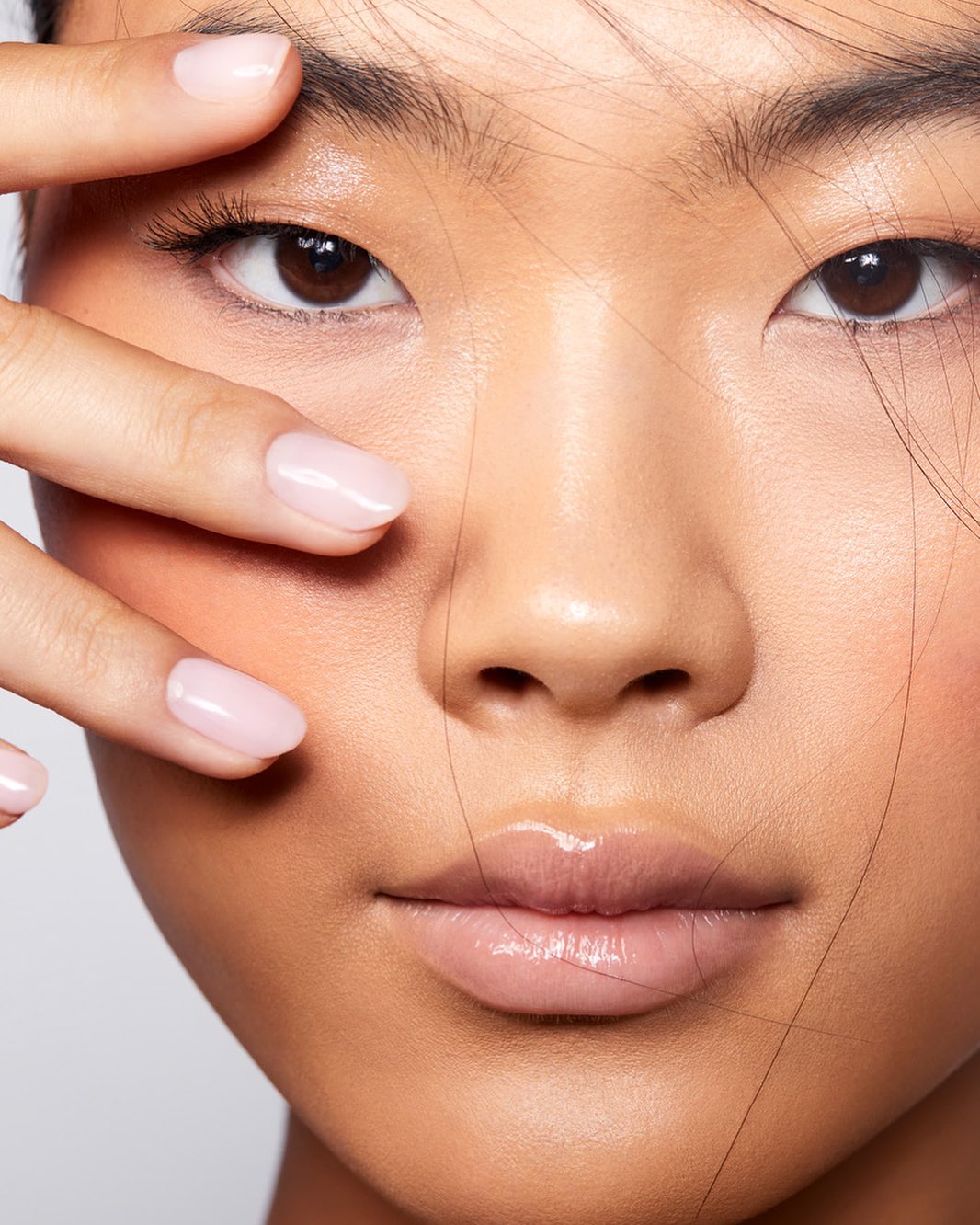 Close-up of an Asian woman's face with glossy lips, soft focus skin, light pink nails, and neutral background. Beauty shot.