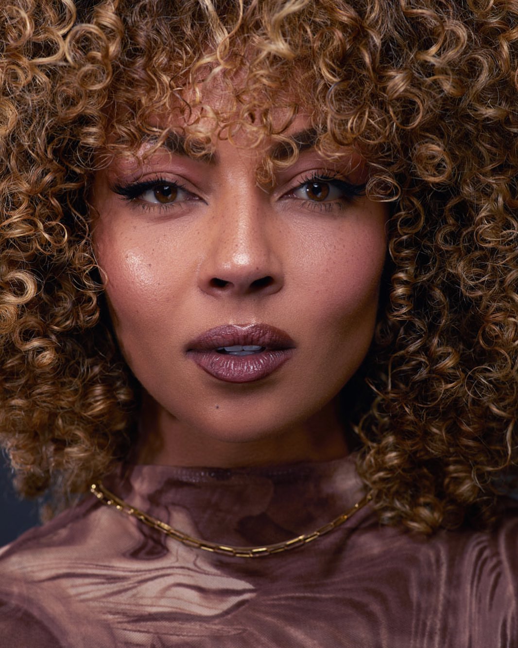 Close-up portrait of a woman with curly hair wearing a patterned top and gold necklace, looking directly at the camera.