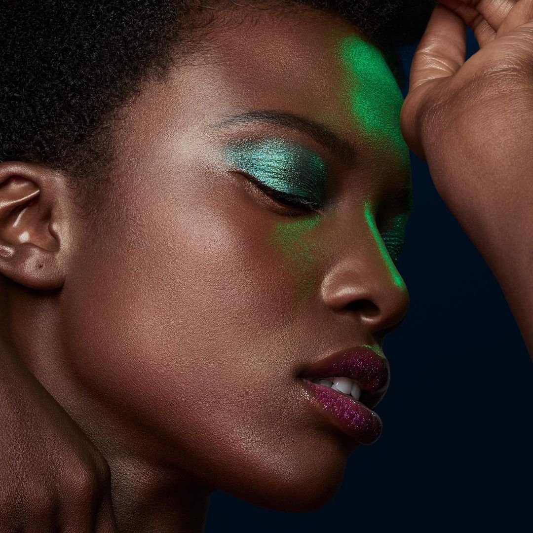 Close-up of a woman with dark skin, iridescent green eyeshadow, and purple glitter lipstick, posing in a dark studio with green lighting.