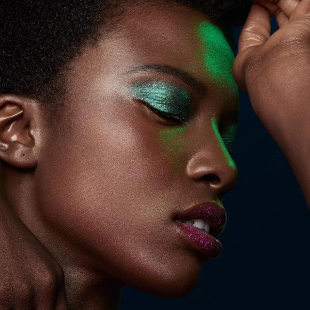 Close-up of a woman with dark skin, iridescent green eyeshadow, and purple glitter lipstick, posing in a dark studio with green lighting.