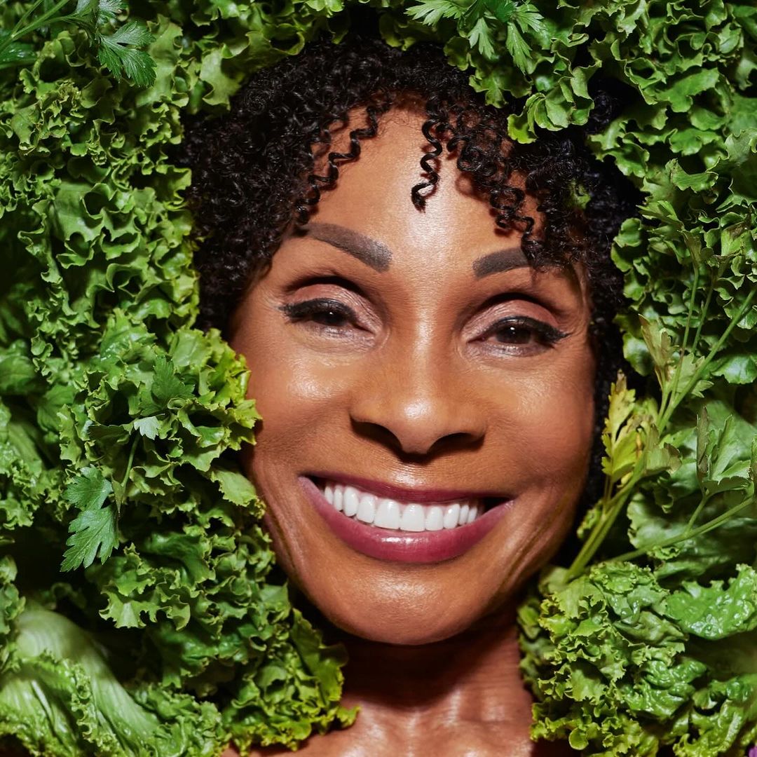 Close-up portrait of a smiling African-American woman surrounded by vibrant green lettuce leaves and sprigs of parsley.