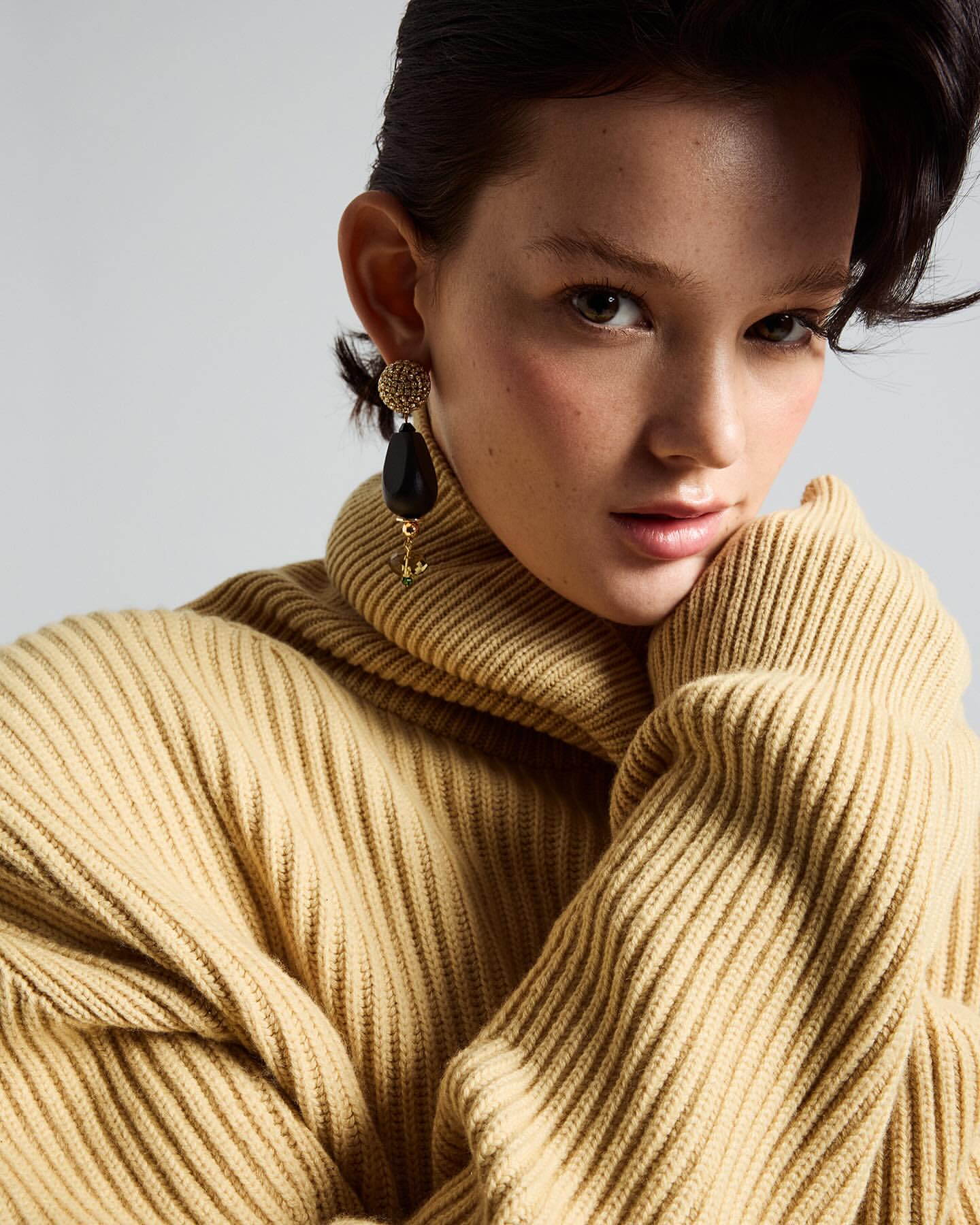 Close-up portrait of a woman wearing a heavy ribbed turtleneck sweater and large drop earrings against a plain background