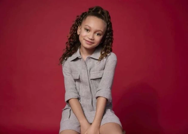 Young girl with curly brown hair wearing a grey utility playsuit against a solid deep red background.