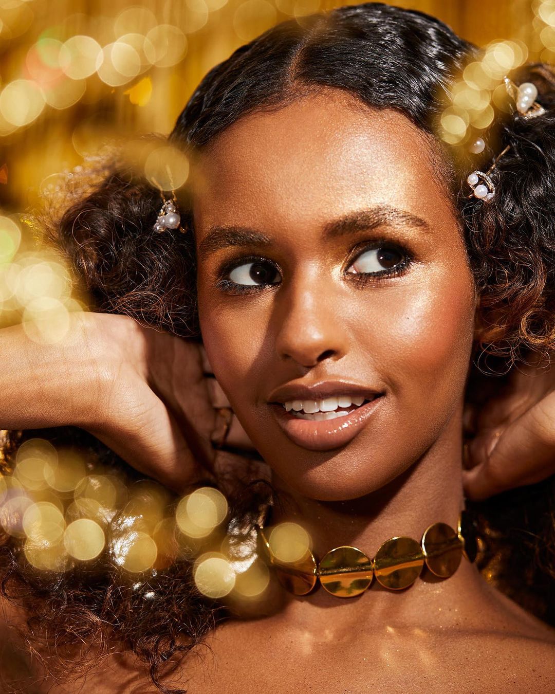 Close-up portrait of a smiling woman with dark curly hair adorned with pearls, wearing a gold choker against a shimmering gold background.
