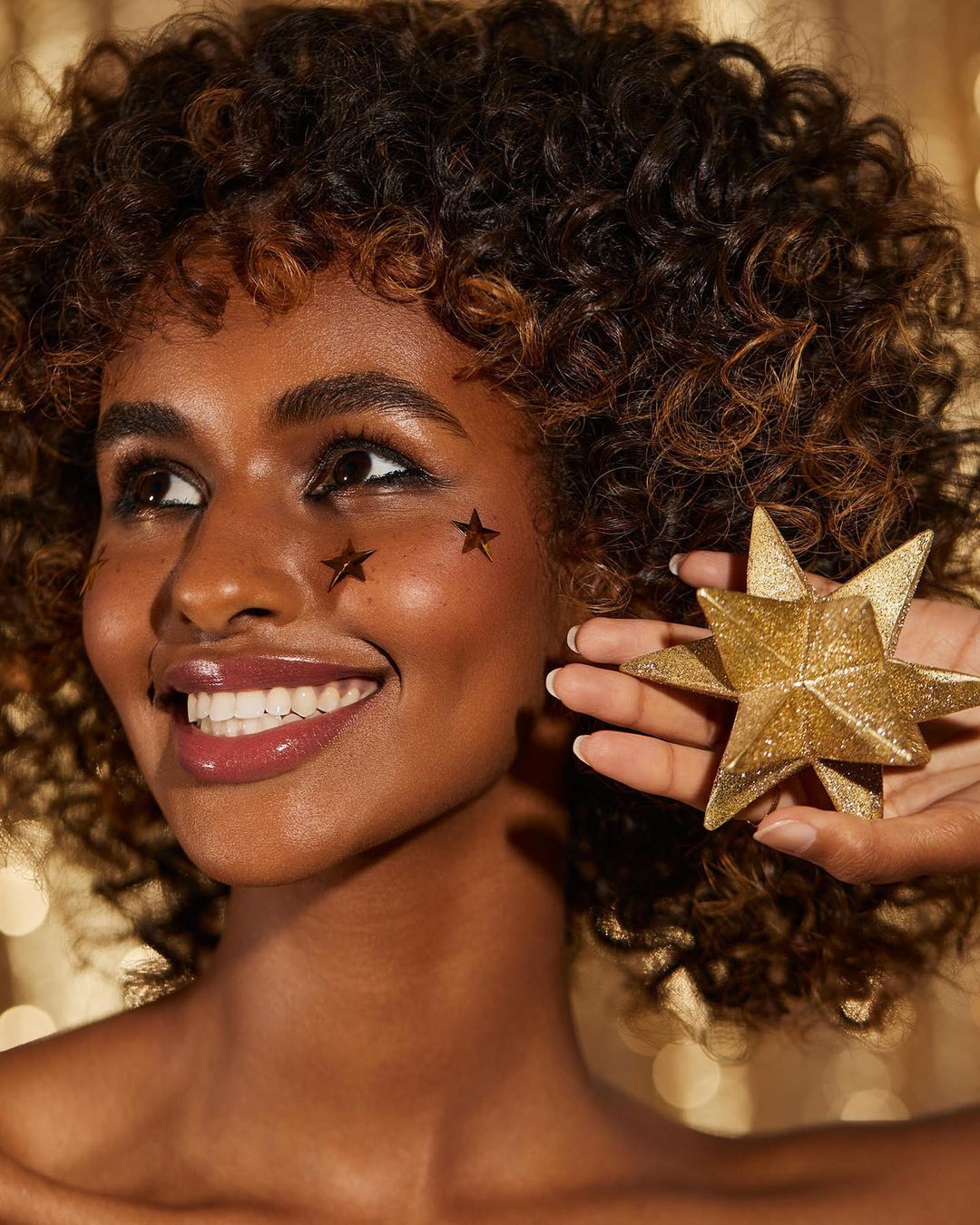 Close-up of smiling woman with curly hair and star decorations on her face, holding a gold star in her hand against bokeh lights.