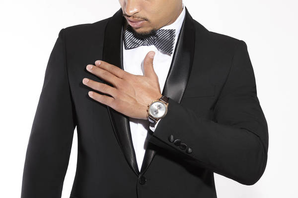 Close-up studio shot of an adult male wearing a black tuxedo with bow tie, placing his hand over the chest with a wristwatch visible.