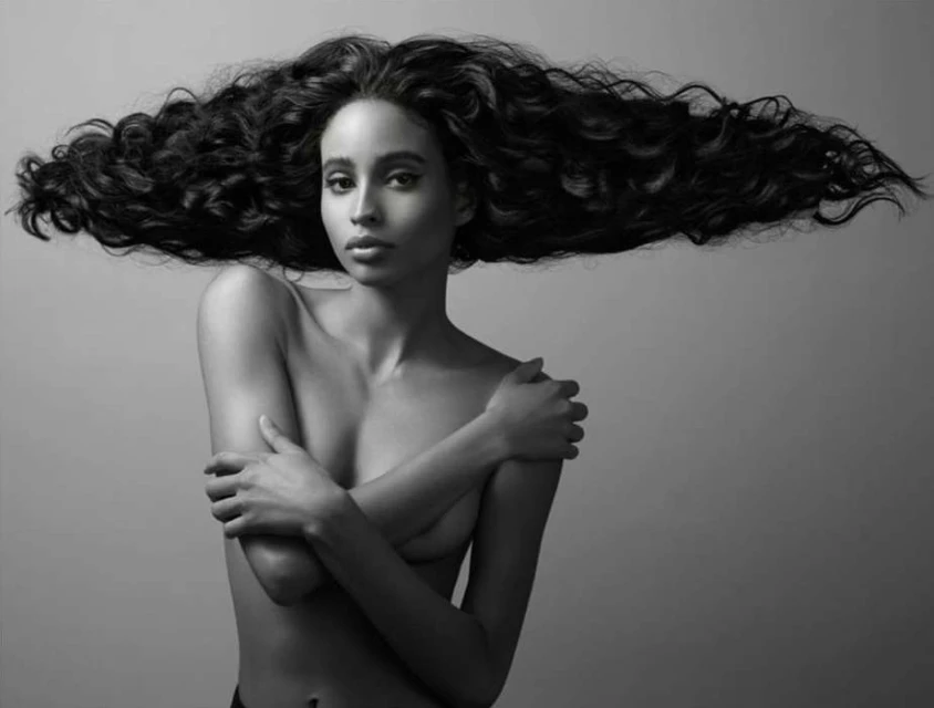 Striking monochrome portrait of a topless woman with flowing, voluminous dark hair against a soft grey backdrop, highlighting beauty and form.