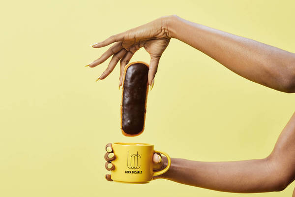 Woman with yellow manicure holds chocolate eclair over a yellow mug against a yellow background in a studio setting.