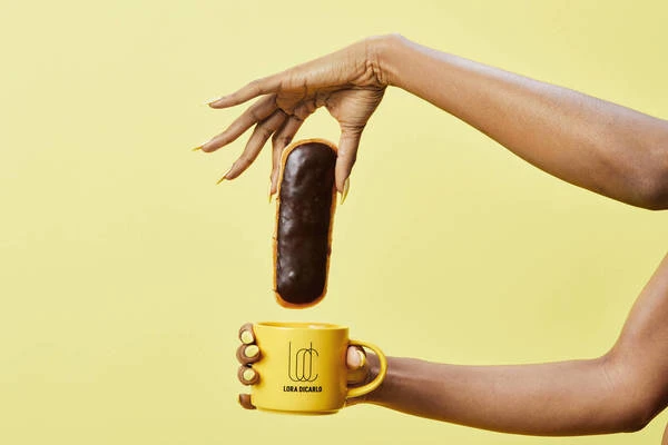 Woman with yellow manicure holds chocolate eclair over a yellow mug against a yellow background in a studio setting.