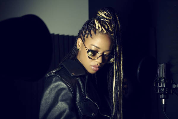 Portrait of an African-American woman with braids, sunglasses, and a leather jacket in a music studio with a microphone.