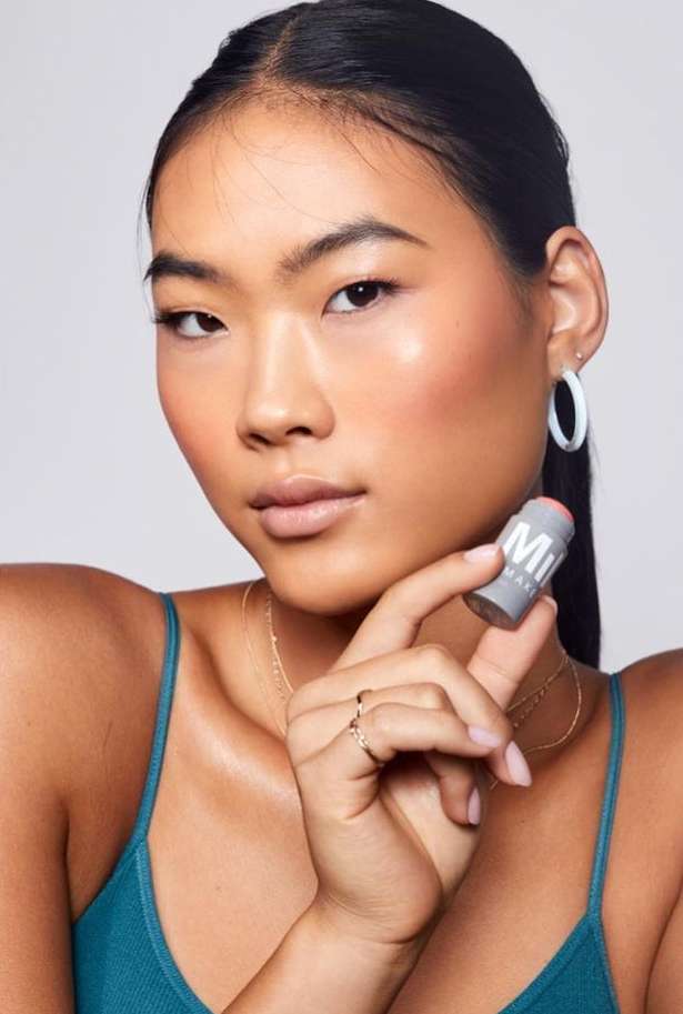 Close-up of a young Asian woman holding a Milk Makeup stick. She has her hair pulled back and is wearing a green top.
