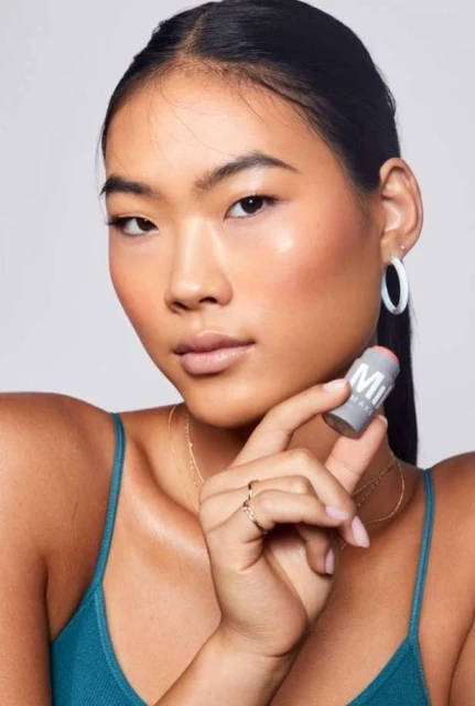 Close-up of a young Asian woman holding a Milk Makeup stick. She has her hair pulled back and is wearing a green top.