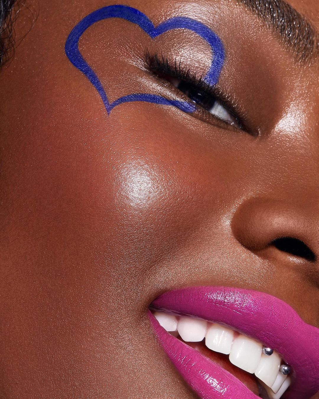 Close-up portrait of an adult female with dark skin, bright pink lips, and a blue heart drawn around her eye.