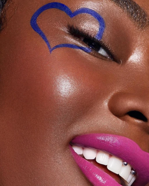 Close-up portrait of an adult female with dark skin, bright pink lips, and a blue heart drawn around her eye.