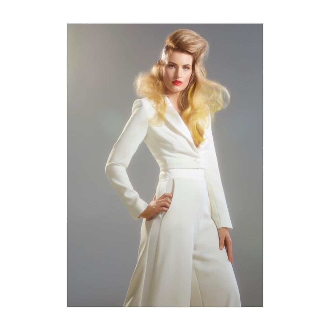 Elegant adult woman in white pantsuit poses in a studio, showcasing blonde hairstyle and red lipstick against a grey background.