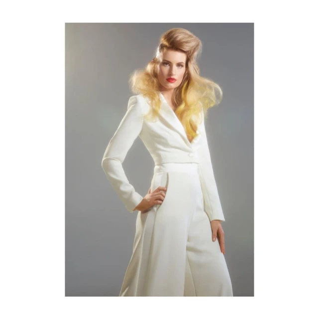 Elegant adult woman in white pantsuit poses in a studio, showcasing blonde hairstyle and red lipstick against a grey background.