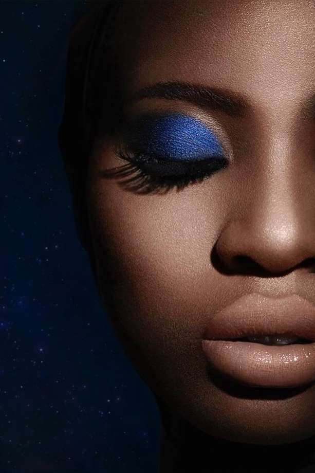 Close-up portrait of a Black woman with vibrant blue eyeshadow and neutral lips against a dark starry background