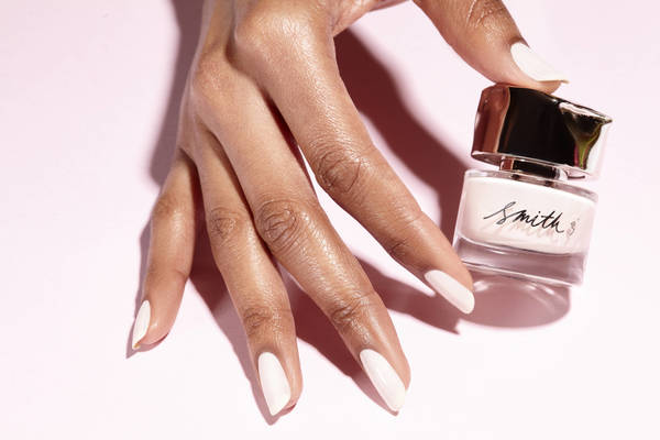 Close-up shot of a dark-skinned hand holding a bottle of Smith & Cult nail polish against a light pink backdrop, showing a beauty product.
