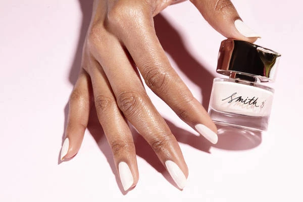 Close-up shot of a dark-skinned hand holding a bottle of Smith & Cult nail polish against a light pink backdrop, showing a beauty product.