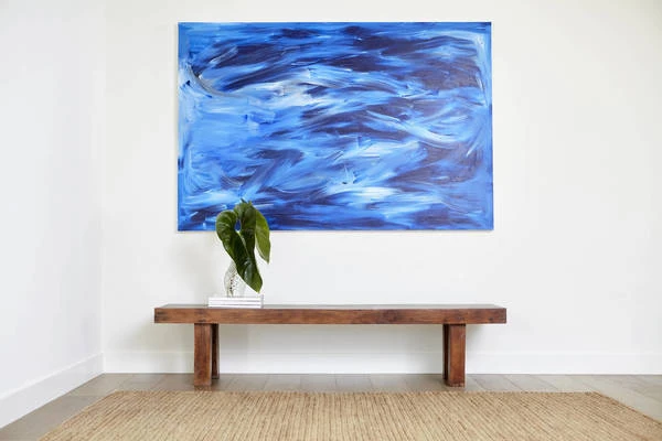 A wooden bench with a vase containing a green leaf sits beneath a large blue abstract painting on a white wall.