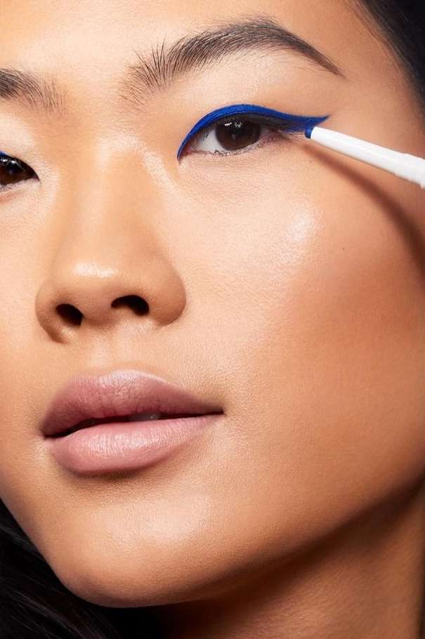 Close-up of an Asian woman applying bright blue eyeliner with a white pencil, highlighting her eye makeup and facial features.