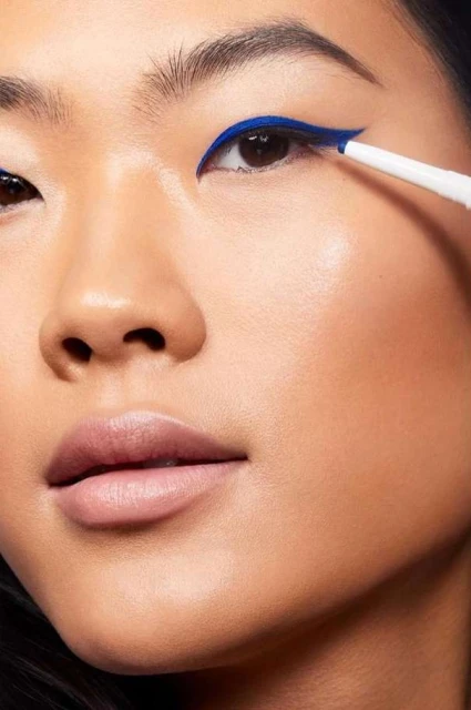 Close-up of an Asian woman applying bright blue eyeliner with a white pencil, highlighting her eye makeup and facial features.