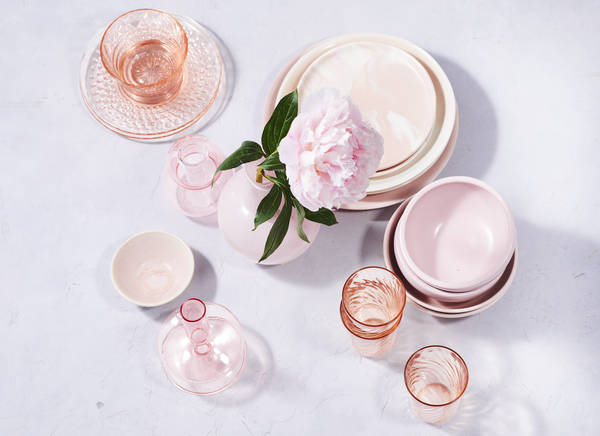 Overhead shot of pink tableware and glassware arranged with a pink peony in a vase on a pale surface, casting soft shadows.