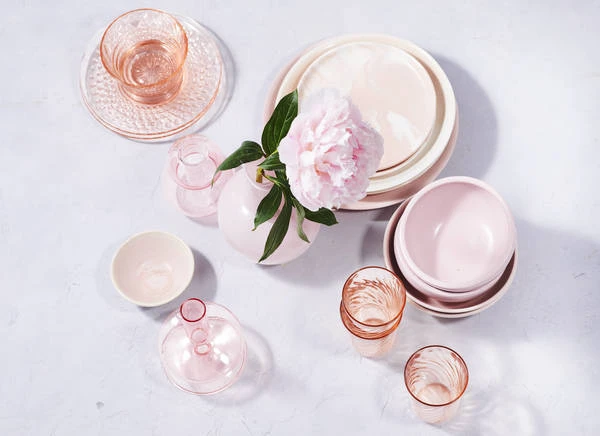 Overhead shot of pink tableware and glassware arranged with a pink peony in a vase on a pale surface, casting soft shadows.
