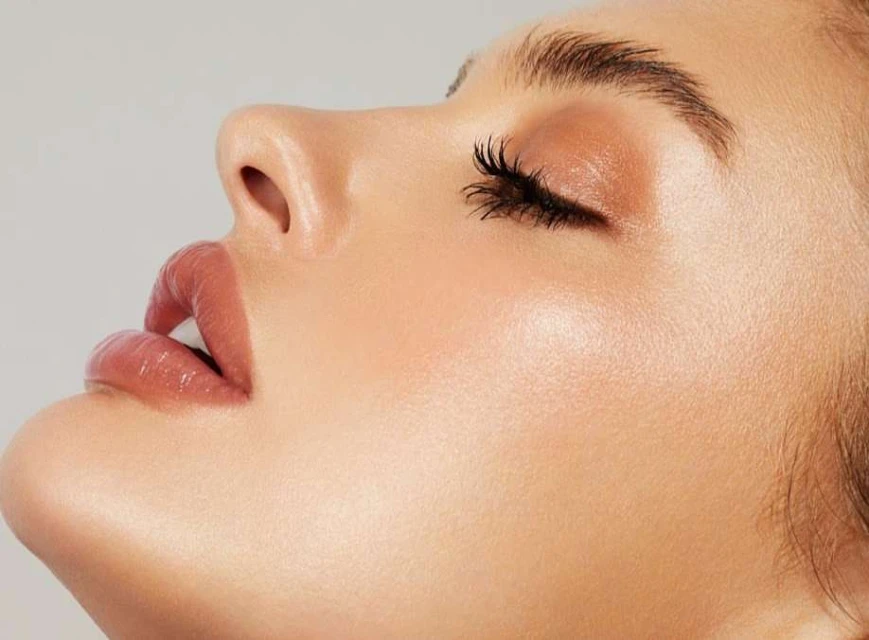 Close-up profile of an adult female's face featuring flawless skin, full lips, long eyelashes, and makeup in a beauty shot against a gray backdrop.
