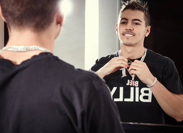Adult male wearing diamond chain and bracelets in front of a mirror reflecting his image and a t-shirt with the text, 'BILLY'.