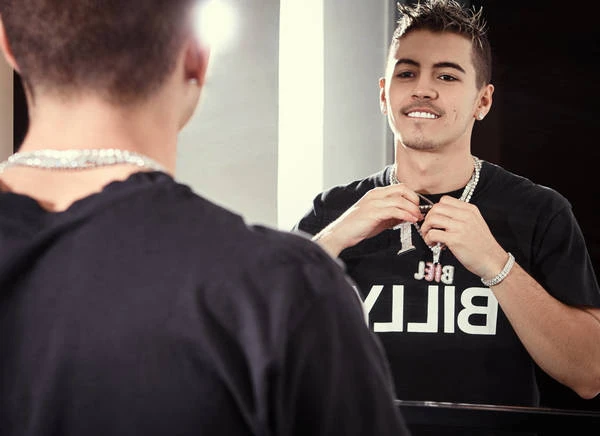 Adult male wearing diamond chain and bracelets in front of a mirror reflecting his image and a t-shirt with the text, 'BILLY'.