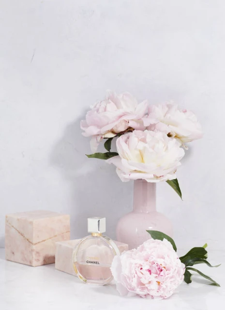 Still life of pink peonies in a vase with Chanel perfume and marble blocks on a white surface against a white background.