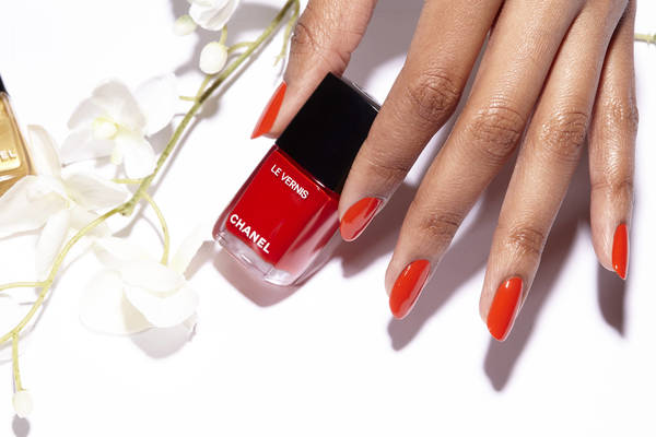Close-up of an adult female's hand holding red Chanel nail polish, showcasing a matching manicure, set against a white background with white flowers.