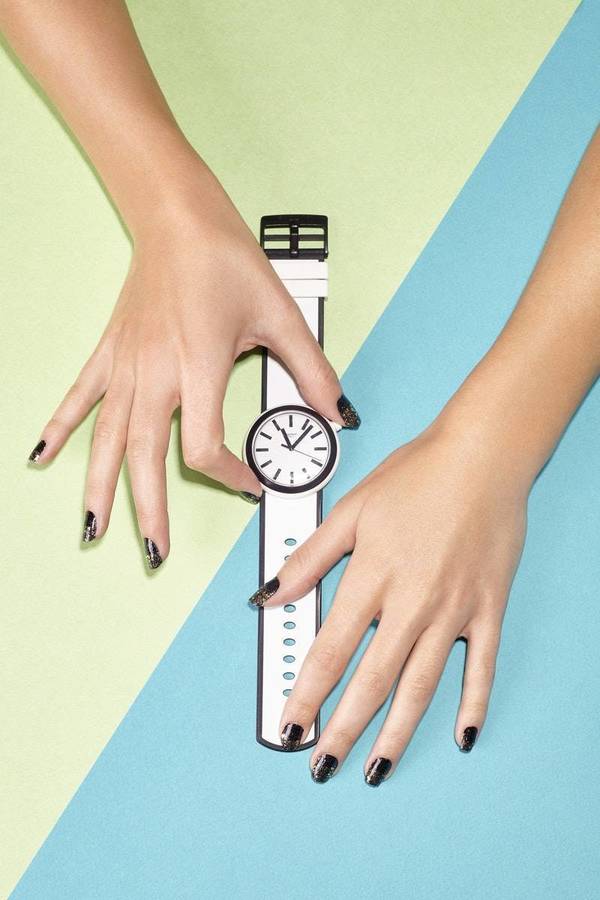 Two hands with dark nail polish holding a white and black wristwatch against a diagonal dual-tone green and blue background.