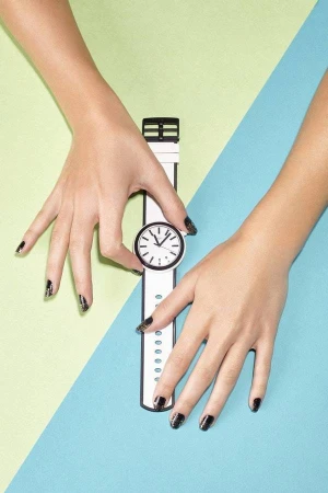 Two hands with dark nail polish holding a white and black wristwatch against a diagonal dual-tone green and blue background.