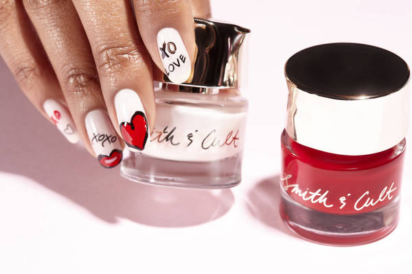Hand with decorated nails holds Smith & Cult nail polish near a red polish on pink backdrop, showcasing beauty and manicured details.