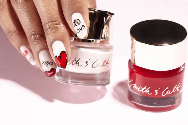 Hand with decorated nails holds Smith & Cult nail polish near a red polish on pink backdrop, showcasing beauty and manicured details.