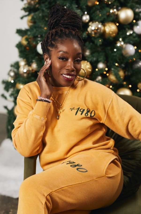 Portrait of a Black woman in a gold-colored tracksuit, seated in front of a Christmas tree decorated with gold and silver ornaments.
