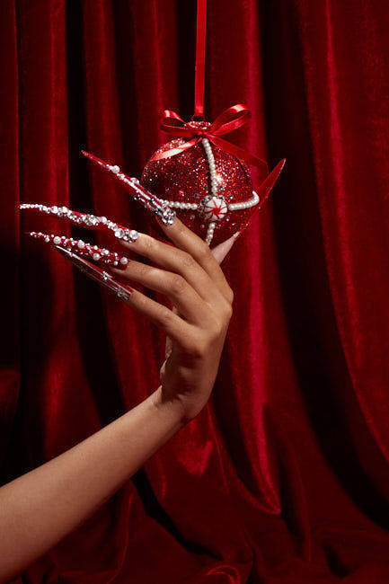 Hand with red, jewel-encrusted long nails holds a matching red sequin Christmas bauble ornament against a dark red velvet curtain.