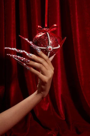 Hand with red, jewel-encrusted long nails holds a matching red sequin Christmas bauble ornament against a dark red velvet curtain.