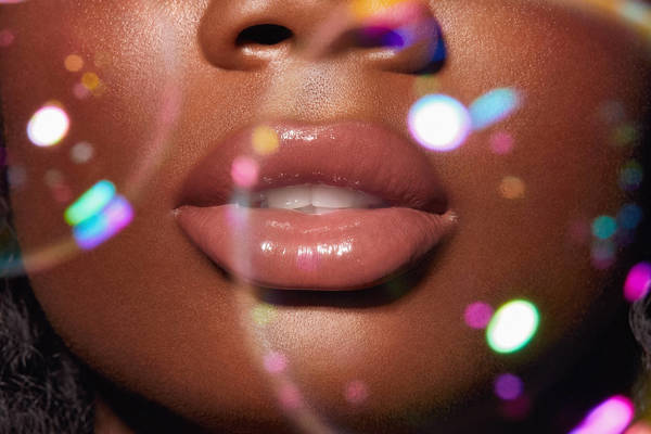 Close-up of a Black woman's face featuring glossy lips, soft skin texture, and colorful bokeh lights creating a glamourous, diffused effect.