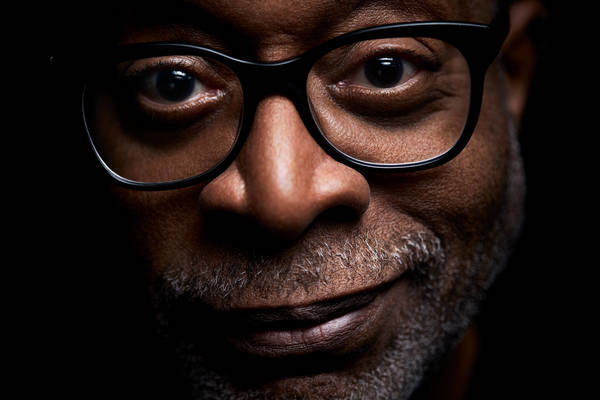 Close-up portrait of an adult male with glasses, a dark complexion, and a hint of a smile, set against a dark background.