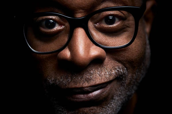 Close-up portrait of an adult male with glasses, a dark complexion, and a hint of a smile, set against a dark background.