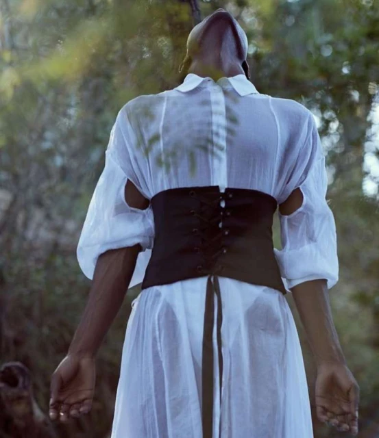 Adult woman in a white dress and black corset stands with head tilted back in a forest environment with blurred tree background.